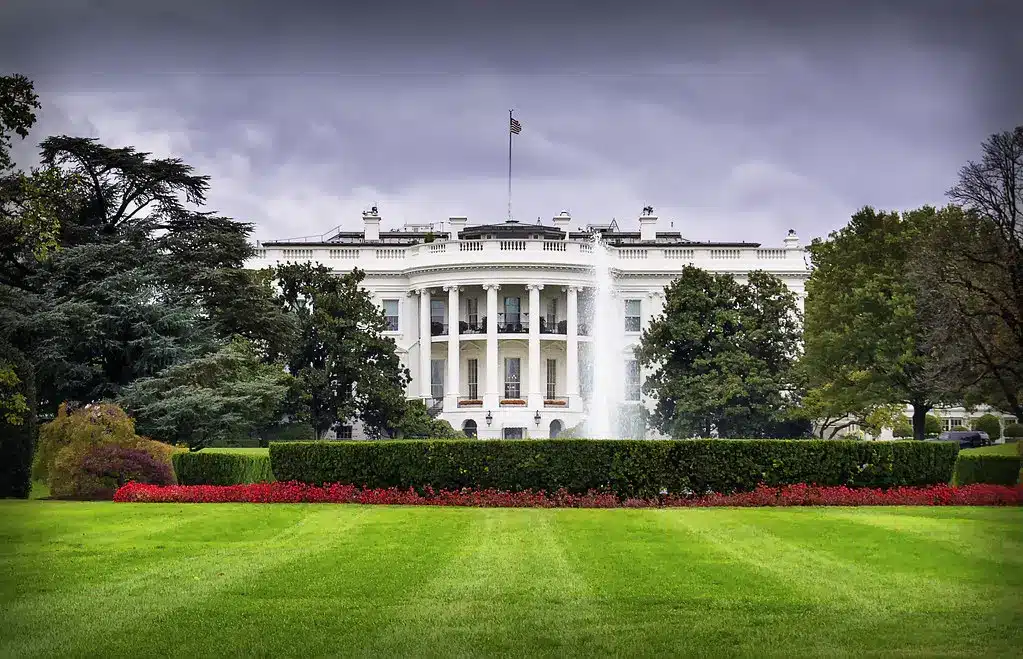 A wide shot of the White House facade behind a manicured green lawn and red flower beds under a cloudy sky.