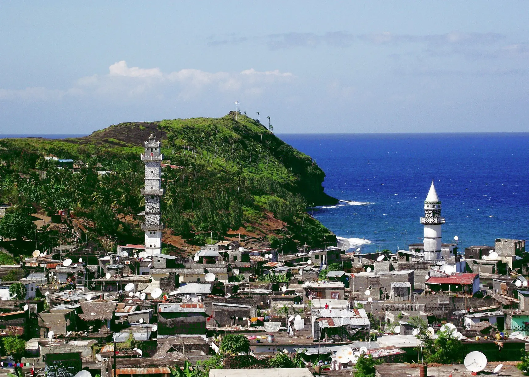 A coastal town in Anjouan with white buildings, minarets, and lush green hills overlooking the blue ocean.